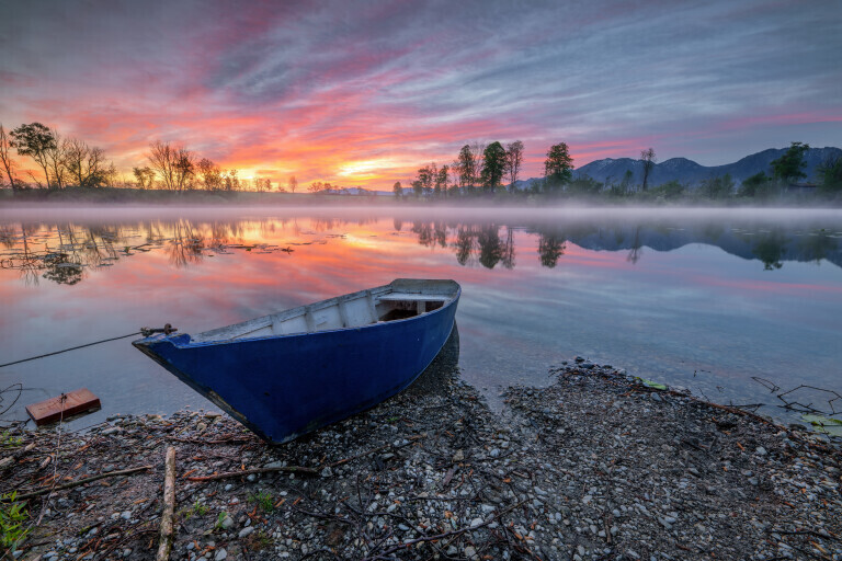 Blaues Boot am See zum Sonnenaufgang mit Bergen und roten WOlken